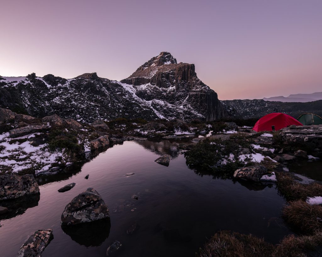 Red tent camped beside reflective alpine tarn at purple dusk, Tasmanian mountains