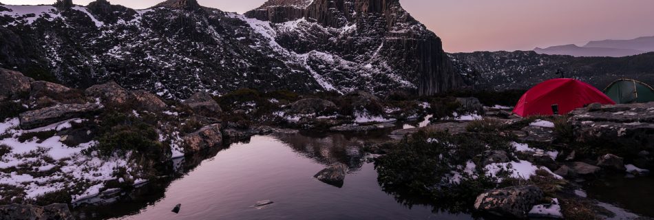 Red tent camped beside reflective alpine tarn at purple dusk, Tasmanian mountains