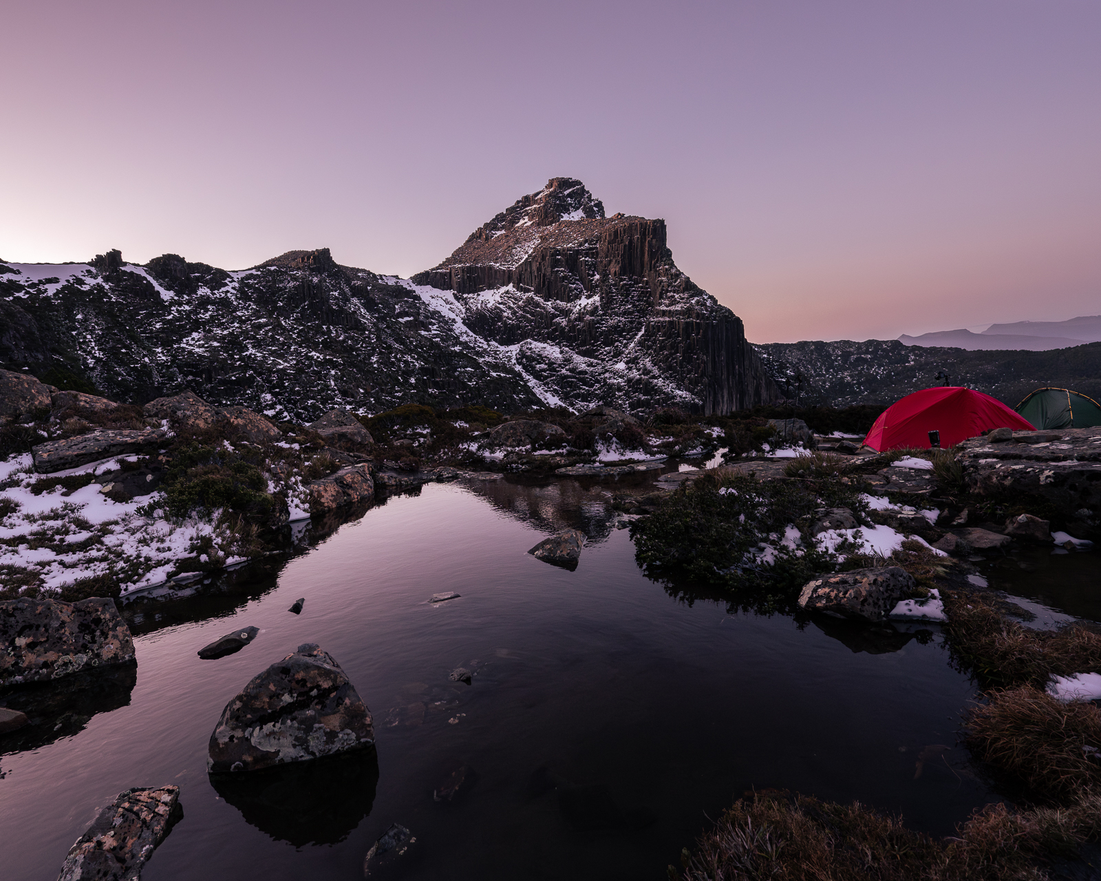 Red tent camped beside reflective alpine tarn at purple dusk, Tasmanian mountains
