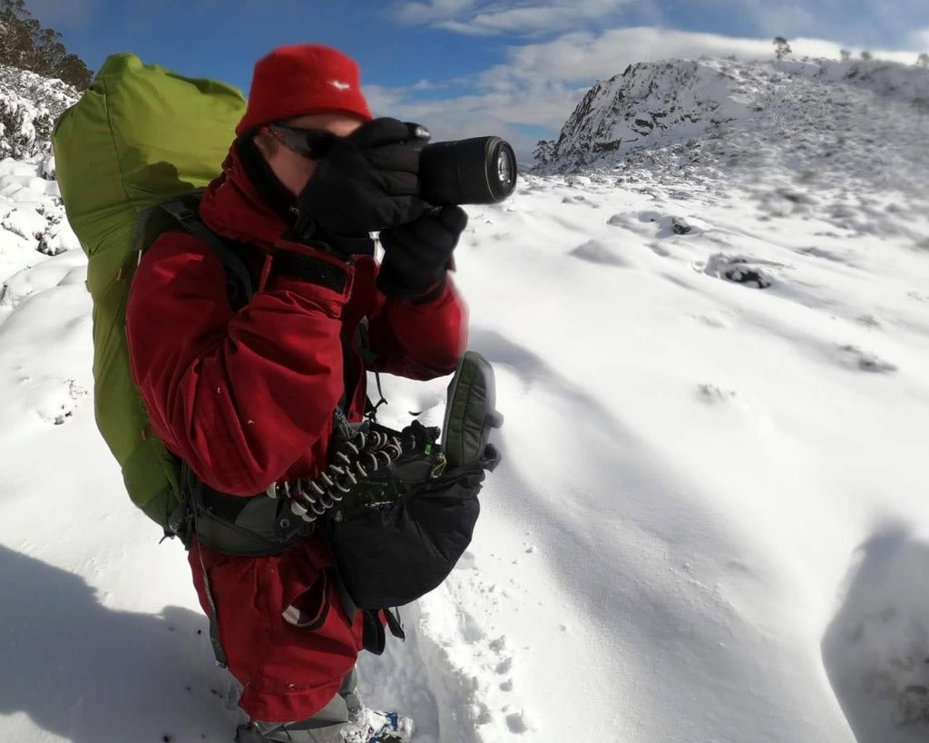 Photographer Francois Fourie in red jacket and beanie photographing snowy Tasmanian alpine landscape with large camera and heavy backpack