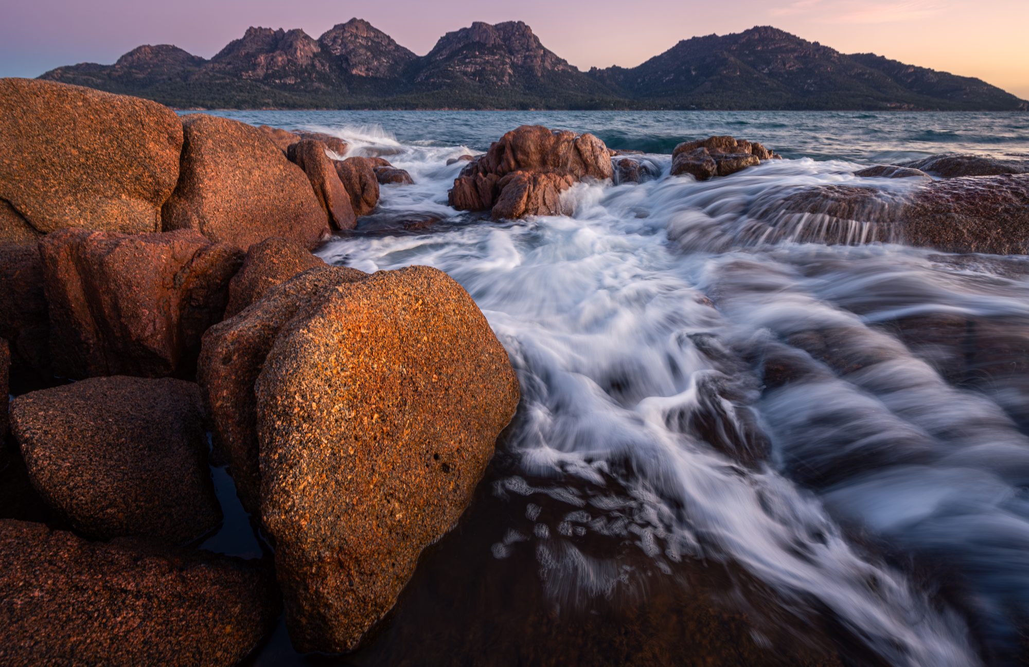Golden sunset light over Coles Bay with pink granite peaks reflected in calm Tasmanian coastal waters