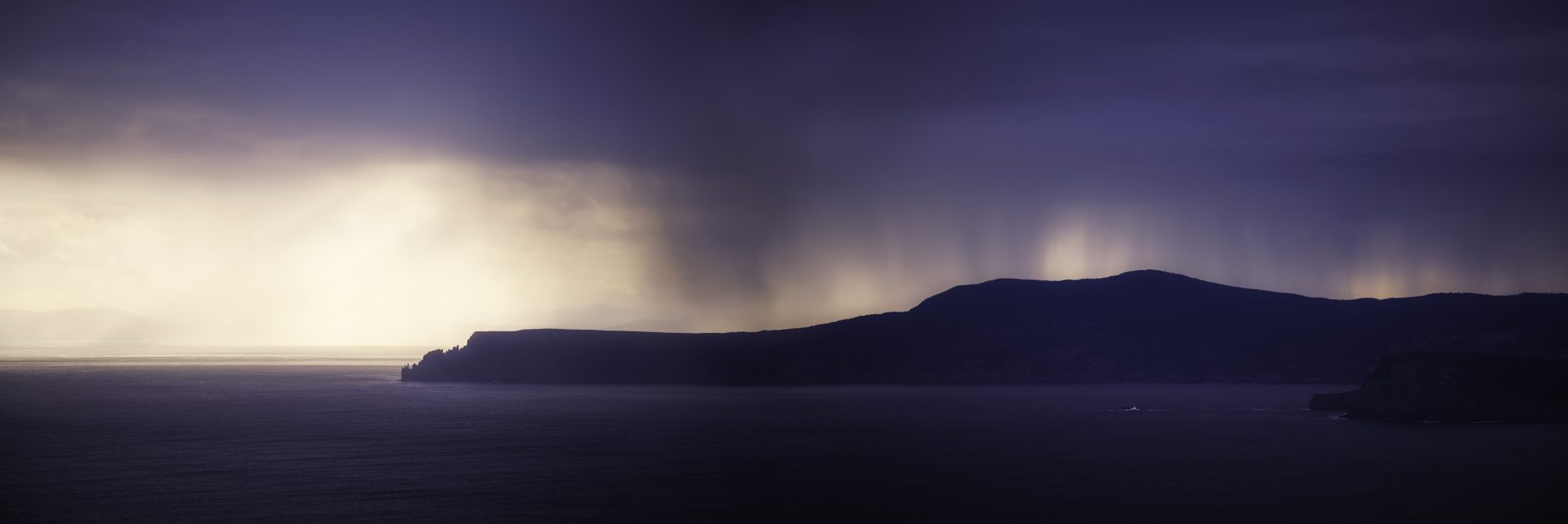 Panoramic view of storm rain curtains and golden sunset light over a dark Tasmanian coastal headland with calm sea in the foreground