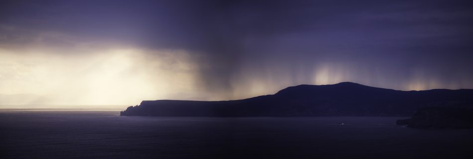 Panoramic view of storm rain curtains and golden sunset light over a dark Tasmanian coastal headland with calm sea in the foreground